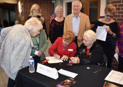 A group of older people standing around a table signing books.