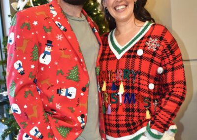 A man and woman wearing ugly christmas sweaters in front of a christmas tree.