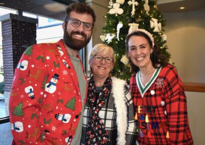 Three people standing in front of a christmas tree wearing ugly sweaters.