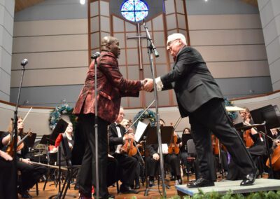 Two men shaking hands in front of an orchestra.